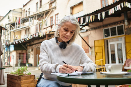 The beautiful gray haired woman sits at a table, writing attentively in a lively courtyard.の写真素材