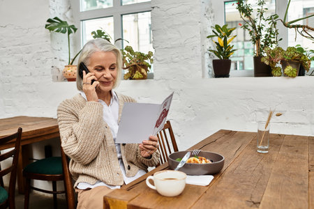 A beautiful gray haired woman sits at a wooden table, enjoying breakfast and making a call.の写真素材
