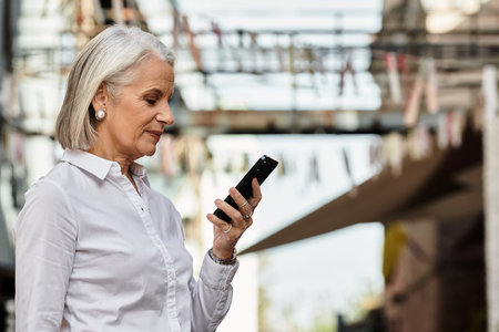 A mature woman admires her smartphone in a vibrant outdoor market filled with colors.の写真素材
