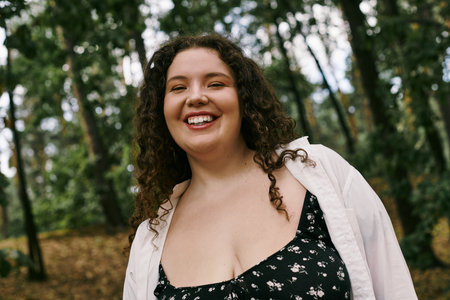 A cheerful plus size woman with curly hair revels in the beauty of nature surrounded by trees.の写真素材