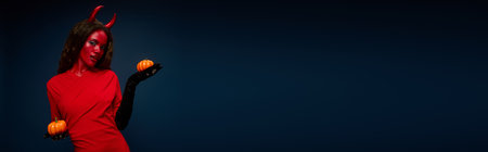 A captivating woman dressed as a devil showcases mini pumpkins with elegance and allure.の写真素材