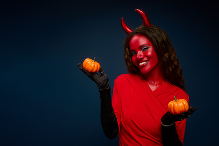 A beautiful woman, dressed as a devil, joyfully holds pumpkins, embracing the Halloween spirit.の写真素材