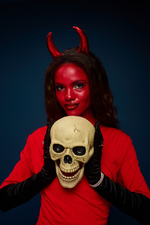 A captivating woman dressed as a devil holds a skull, showcasing Halloween spirit and creativity.