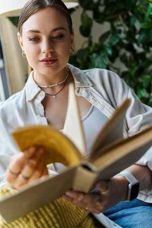 A young woman in casual attire reads a book while sitting comfortably in her home workspace.