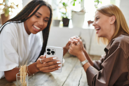 Two women relax at a cafe, sharing a joyful moment as they look at a phone together, smiling brightly.の写真素材