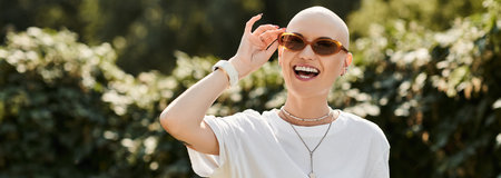 A young bald woman in elegant attire playfully adjusts her sunglasses while smiling outdoors.の写真素材