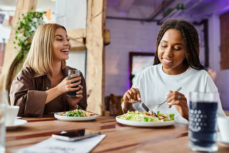 A smiling woman with braided hair enjoys a meal while her girlfriend looks on with delight at a cafe.の写真素材