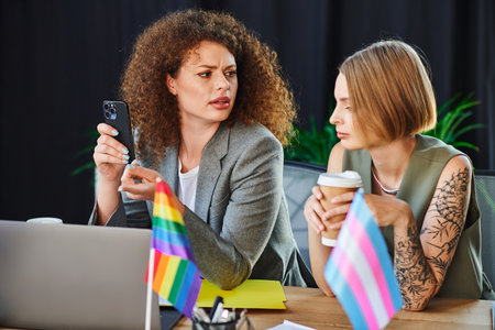 Two coworkers share a thoughtful conversation while surrounded by pride flags and office materials.の写真素材