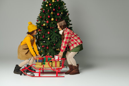 girls excitedly push a sled filled with colorful presents near a decorated Christmas tree.の写真素材