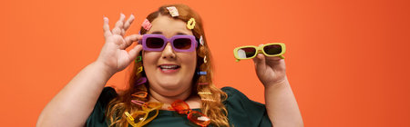 A cheerful plus size woman with stylish clips in her hair displays two pairs of trendy sunglasses.