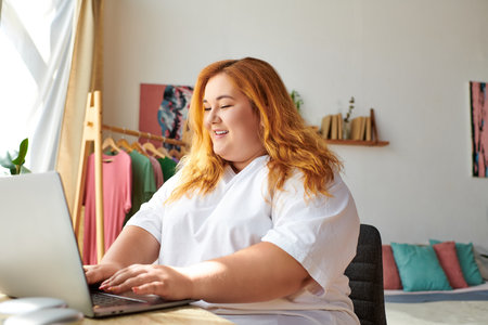 The plus size woman enjoys her time typing on the laptop, surrounded by a cheerful setting.の写真素材