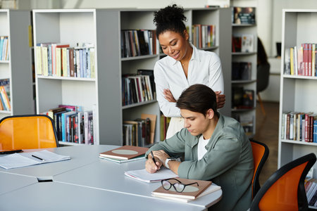 A passionate educator offers support to her student engaged in his studies at a library.