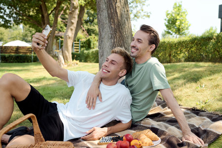 Two men enjoy a delightful picnic, smiling while taking a selfie together in a serene park.の写真素材