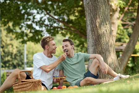 Two happy men enjoy a lovely picnic, feeding each other under a tree on a warm day.の写真素材