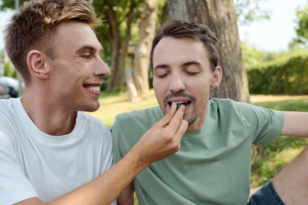 Two smiling men enjoy a playful moment while feeding each other outdoors in a beautiful setting.の写真素材
