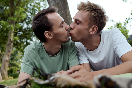 Two men share a tender kiss while lying on the grass, surrounded by natures beauty.の写真素材