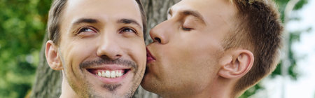 Two joyful men share an affectionate kiss surrounded by vibrant nature, radiating happiness.の写真素材