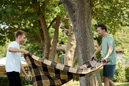 Two men laugh and share a lighthearted moment outdoors while handling a blanket under a tree.の写真素材