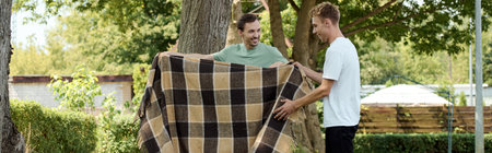 Two smiling men share a light moment as they arrange a blanket in a beautiful outdoor setting.の写真素材