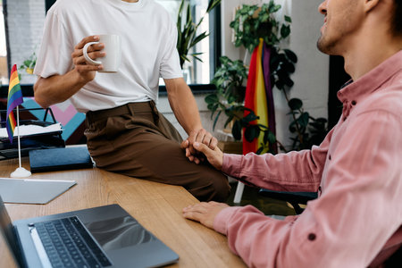 A loving couple shares a tender moment in a modern workspace filled with plants and pride flags.