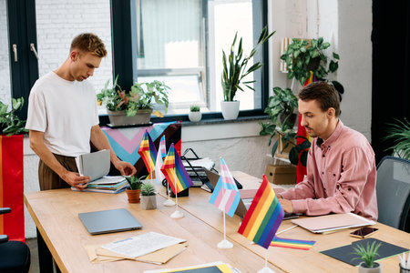 Two men sort paperwork in a lively, plant filled space adorned with pride flags.の写真素材
