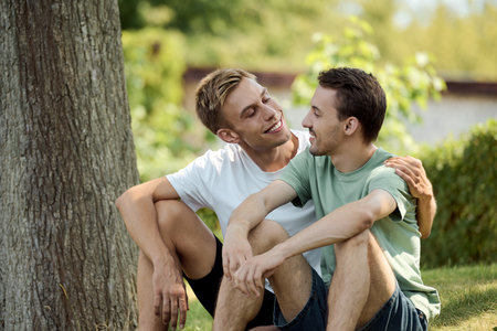 Two smiling men sit close together in a lush park, sharing laughter and affection.の写真素材