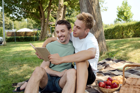 The couple shares an intimate moment, reading together and laughing under a tree on a sunny day.の写真素材