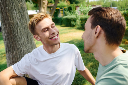 A joyful gay couple sits together in a lush park, sharing smiles and warmth in the sunlight.