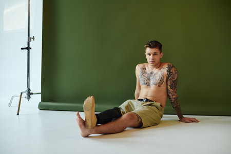 A tattooed young man relaxes in a studio, confidently displaying his prosthetic leg and body art.