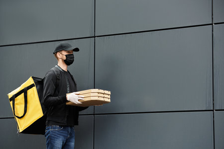 A delivery man wearing a black medical mask brings food orders into an office building while wearing gloves.の写真素材