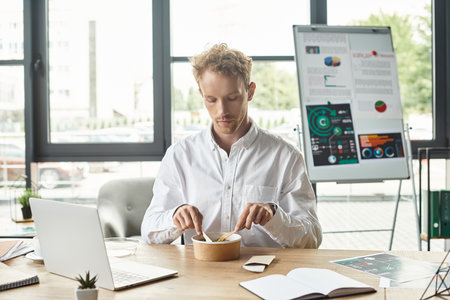 A determined redhead businessman in a white shirt works on a project while enjoying lunch at his...