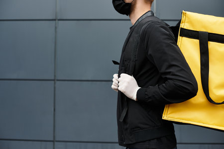 A masked delivery person arrives at an office building, carrying a bright yellow bag filled with...