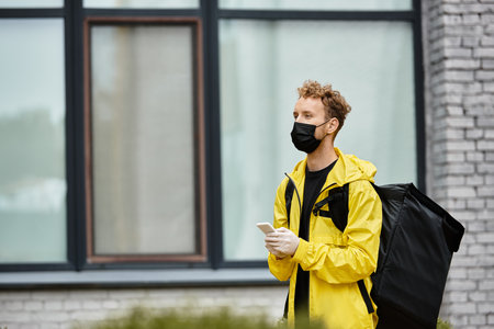 A delivery man wearing a black medical mask stands outside an office building while checking his phone.の写真素材