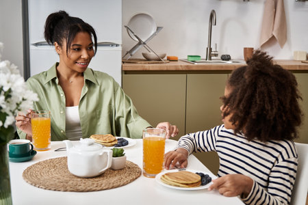 A mother and daughter enjoy a joyful breakfast with pancakes and drinks in their inviting kitchen.の写真素材