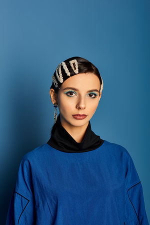 A young woman showcases her trendy attire and accessories in a vibrant blue studio setting.
