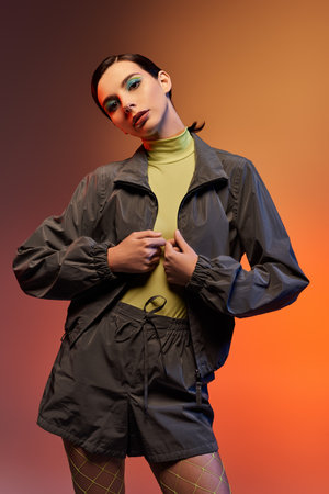 A fashionable young woman poses confidently in a chic outfit against a vibrant orange backdrop.