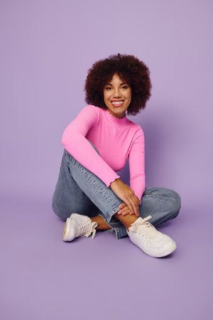 A cheerful young African American woman in pink casual wear sits gracefully with a joyful...
