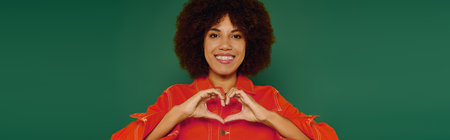A cheerful young woman with curly hair forms a heart shape with her hands, radiating positivity on a green backdrop.の写真素材