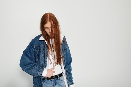 A teenage boy with long red hair confidently poses in a stylish denim outfit against a simple...