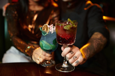 Tattooed couple clinks colorful drinks together, embracing a fun evening at a lively bar.の写真素材