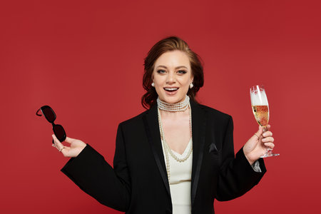 A joyful young woman in stylish attire raises a champagne glass, flaunting her sunglasses.