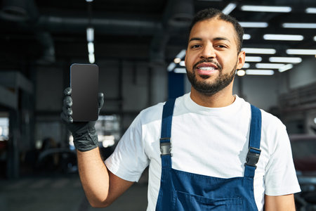 Handsome young mechanic smiles while showcasing a smartphone in a bright workshop setting.の写真素材
