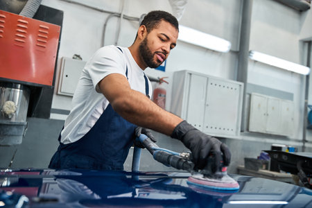 Handsome young mechanic focuses on polishing a vehicle with precision in a busy workshop.