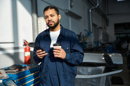 A handsome young mechanic takes a moment to relax with a coffee and phone in the garage.の写真素材