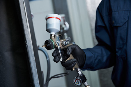 A skilled young mechanic applies paint using a spray gun while working in a bright garage.