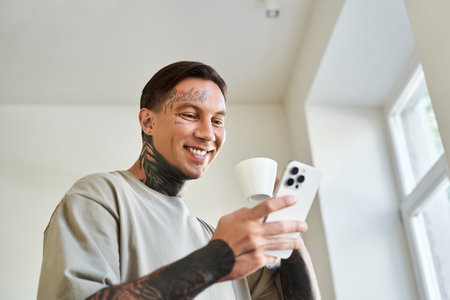A handsome young man smiles as he checks his smartphone with a cup of coffee in hand at home.の写真素材