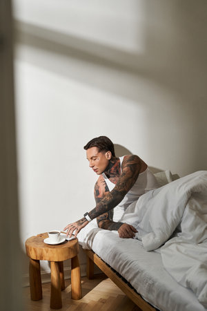 In a serene home setting, a young man reaches for his warm drink beside his cozy bed.の写真素材