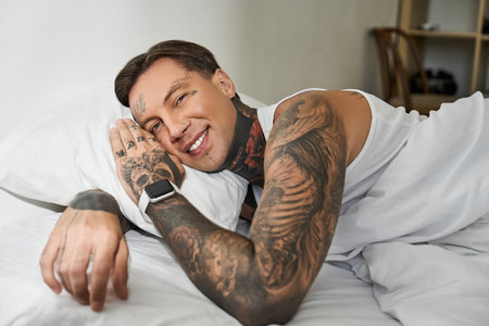 A charming young man with tattoos smiles while lying comfortably on his bed at home.の写真素材