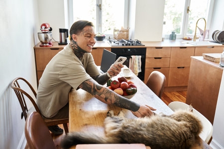 A young man smiles as he sits at the kitchen table, enjoying time with his cat and phone.の写真素材