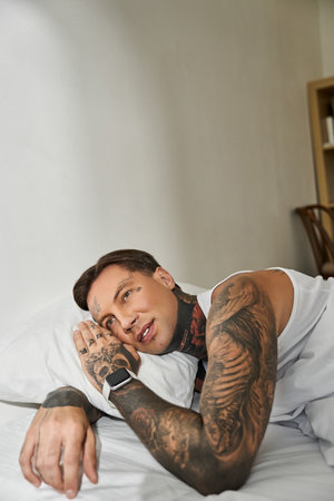 A young man with tattoos lies on a bed, gazing thoughtfully in his serene home environment.の写真素材